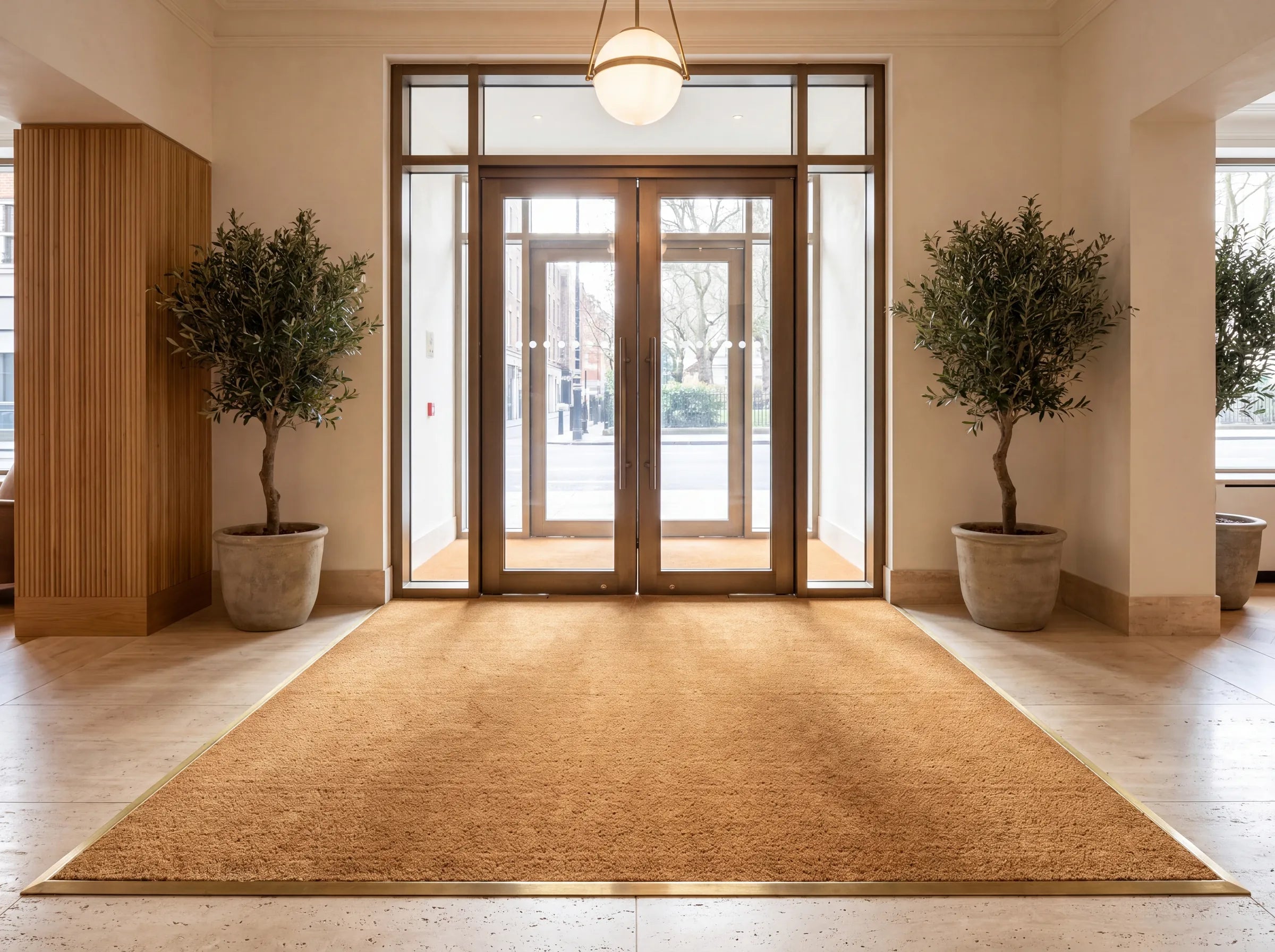 A wall-to-wall custom-cut coir mat set flush in a brass-edged well inside the glass entrance doors of a hotel lobby, between two olive trees